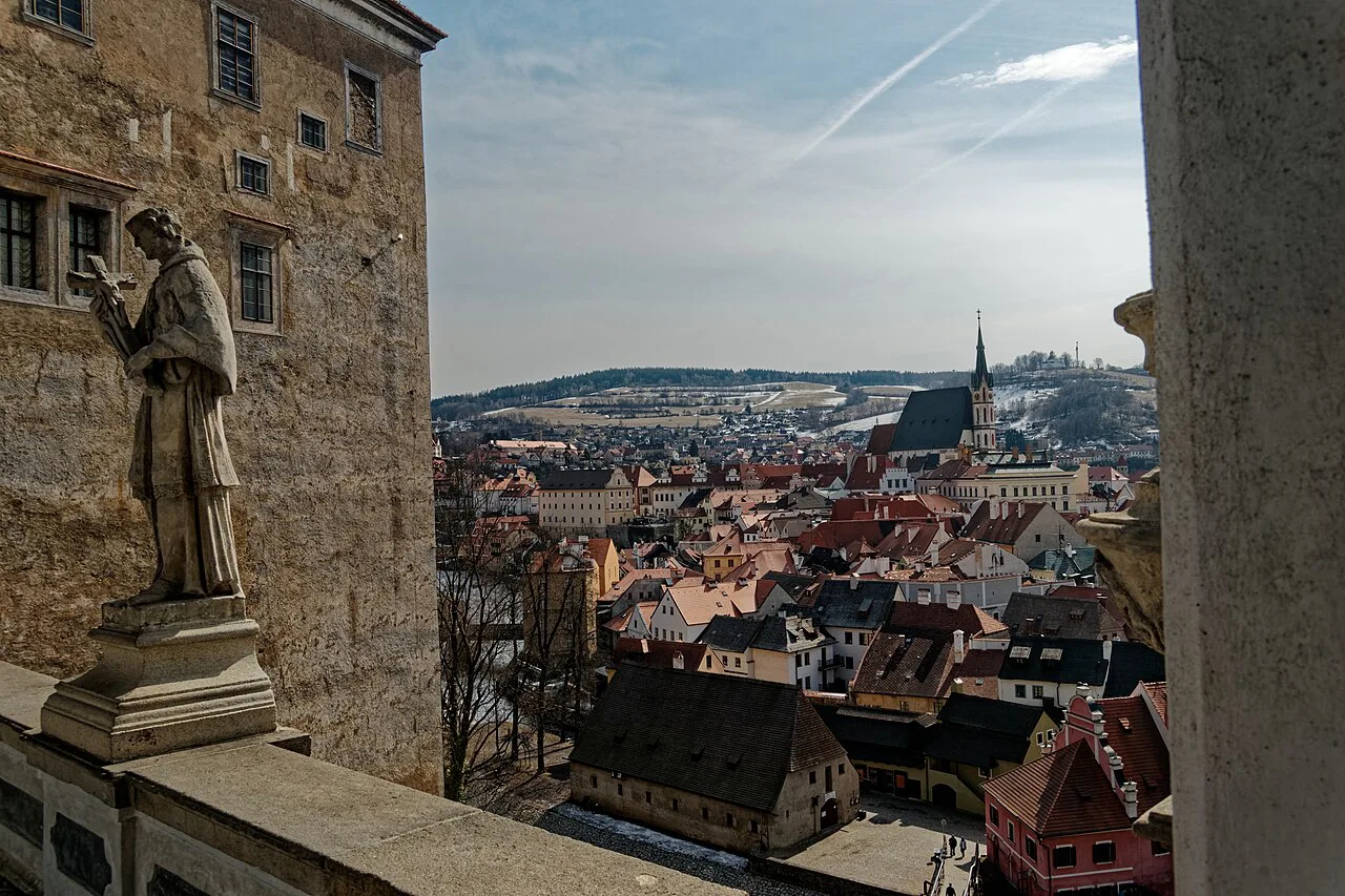 Český Krumlov's rooftops seen from the Cloak Bridge, with a Baroque statue in the foreground.